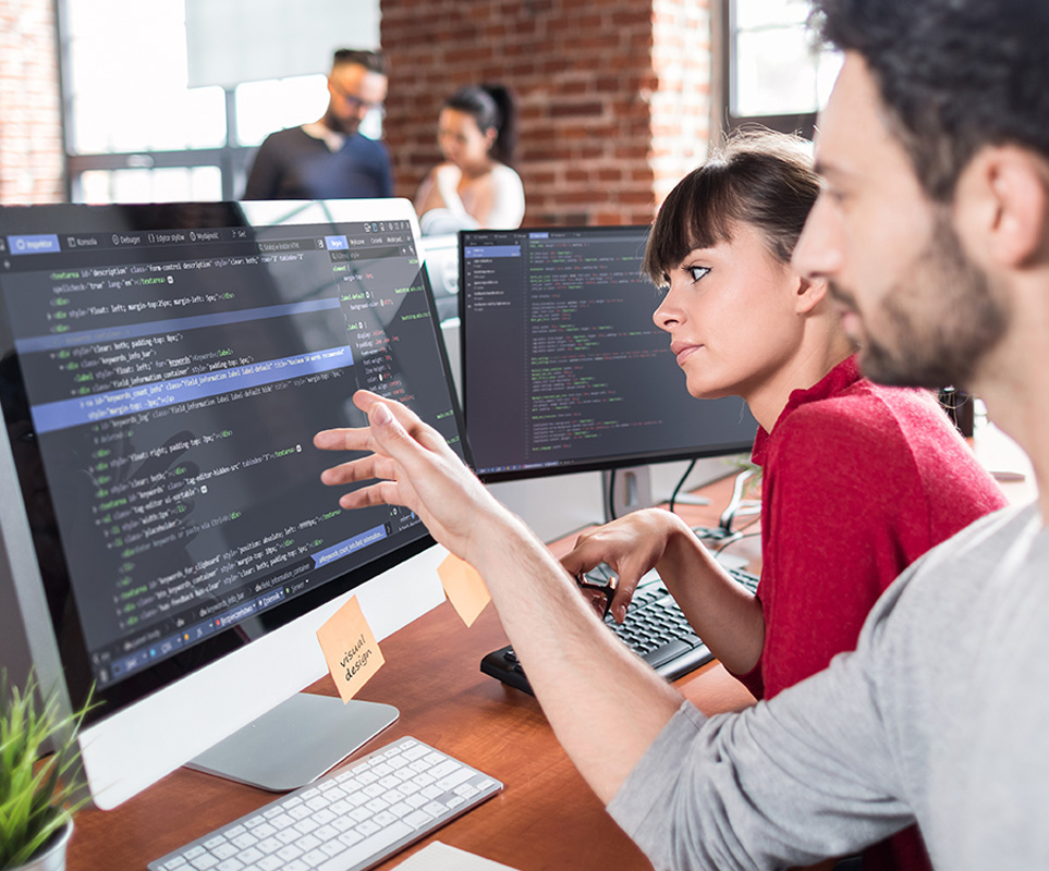 Person pointing at code on a computer screen while discussing it with another, collaborating on CSQE training.